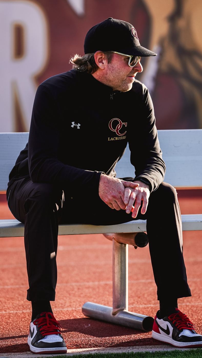 Sean Lindsay today as Oaks Christian head lacrosse coach, sitting on a sideline bench in a black team quarter-zip, cap and sunglasses